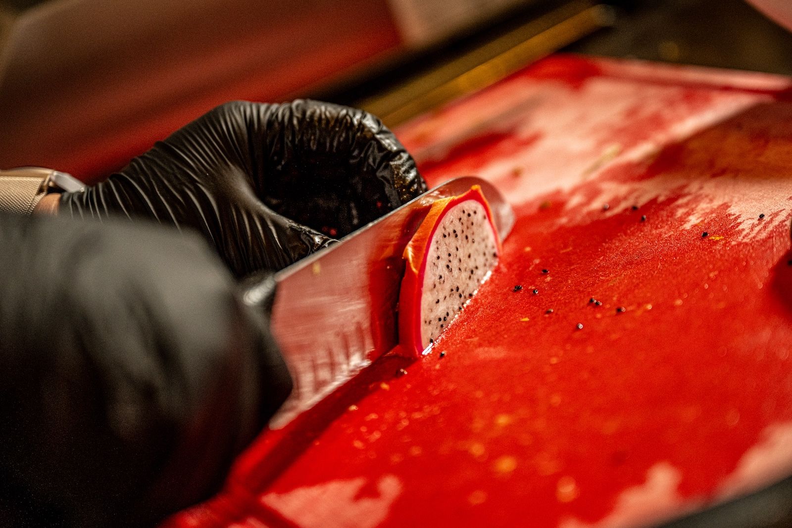 Ein Koch mit schwarzen Handschuhen schneidet sorgfältig eine Drachenfrucht auf einem roten Schneidebrett, während der Saft frisch austritt.