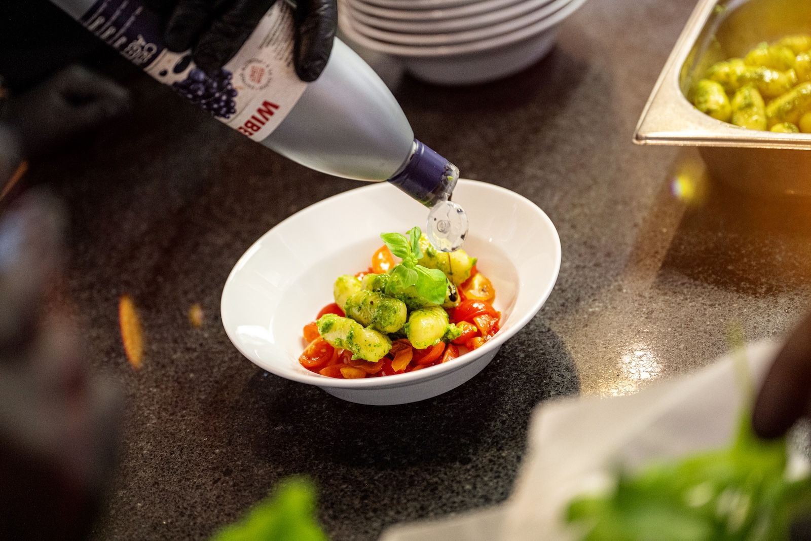 Ein Koch verfeinert frische Gnocchi mit Kirschtomaten und Basilikum in einer weißen Schale, während er vorsichtig Olivenöl darüberträufelt.